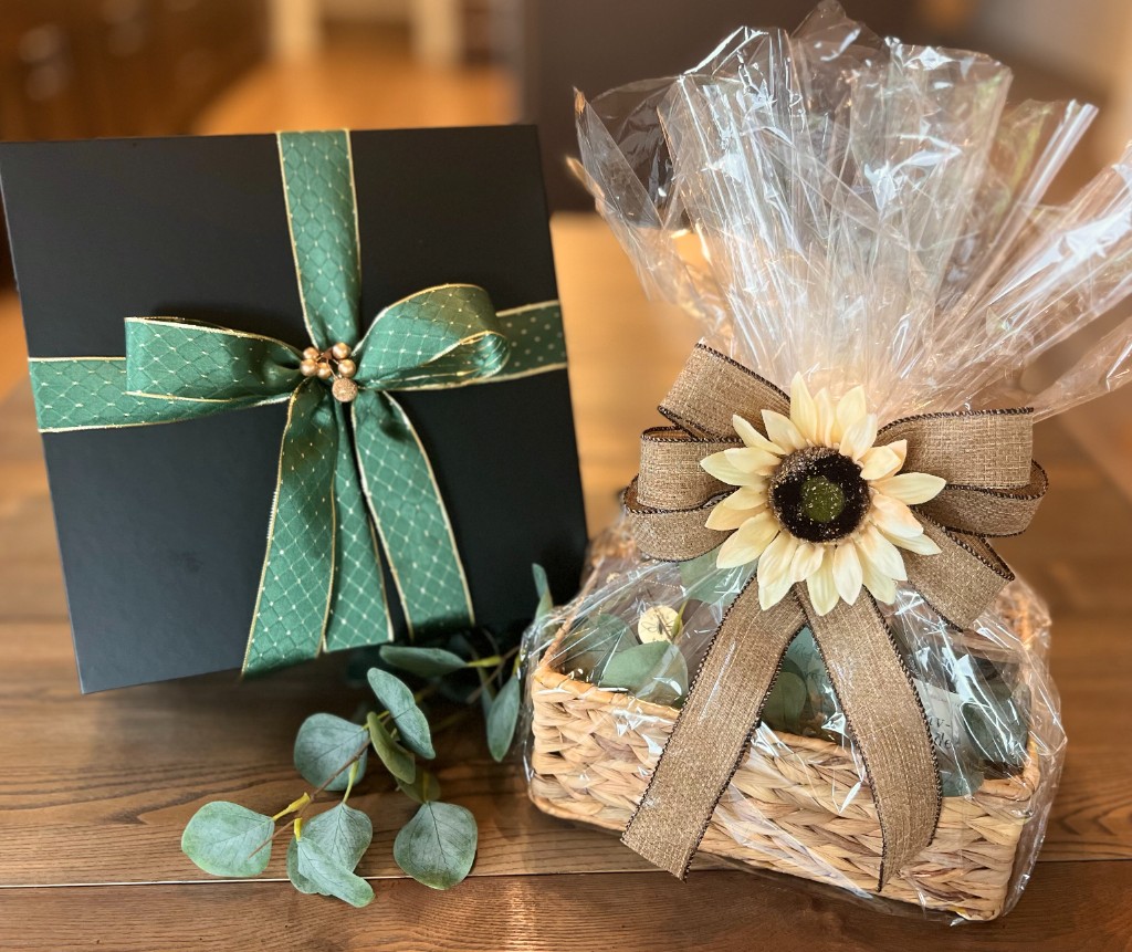 A black gift box with a green ribbon beside a cellophane-wrapped gift basket