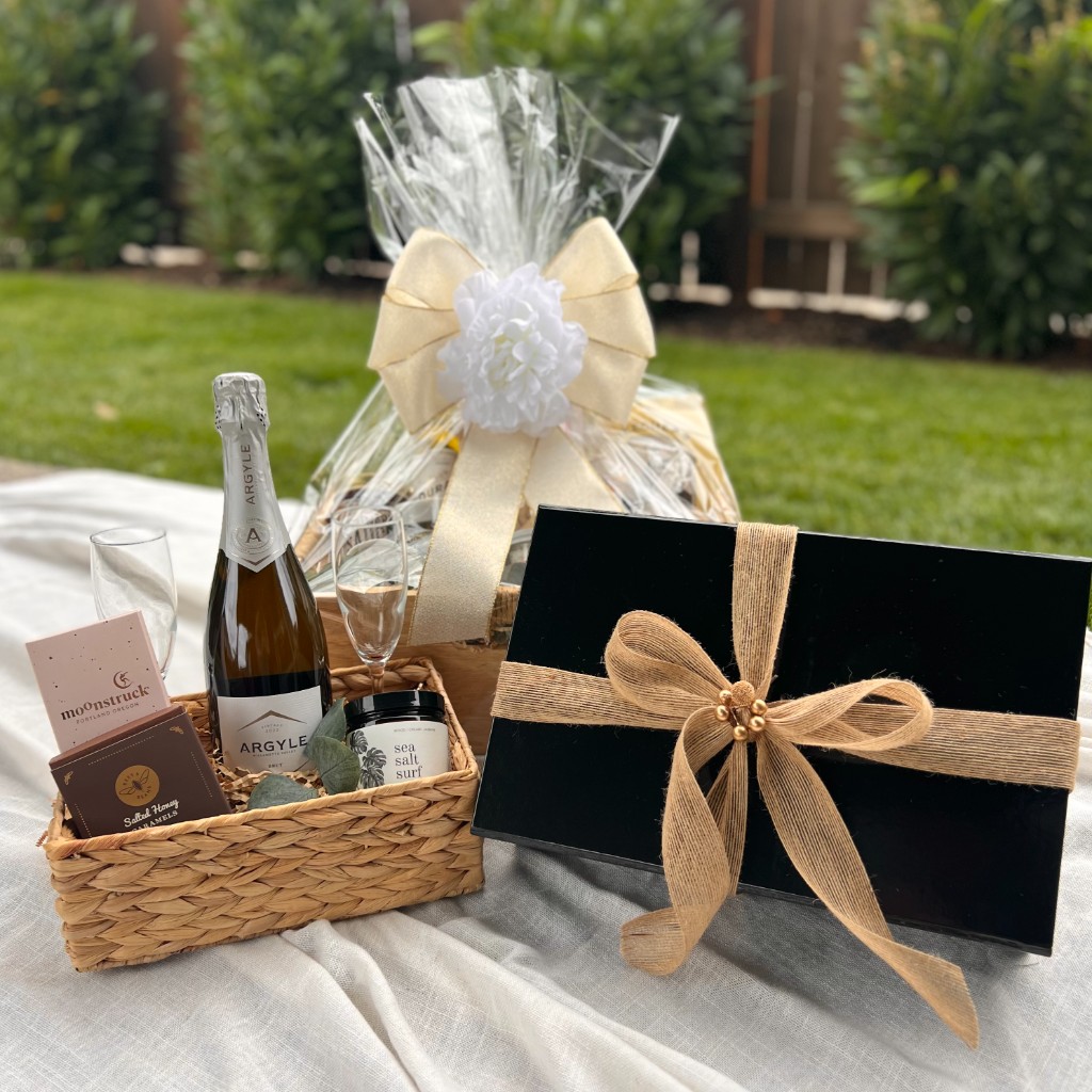 Two gift baskets and a black gift box arranged outdoors on a white cloth